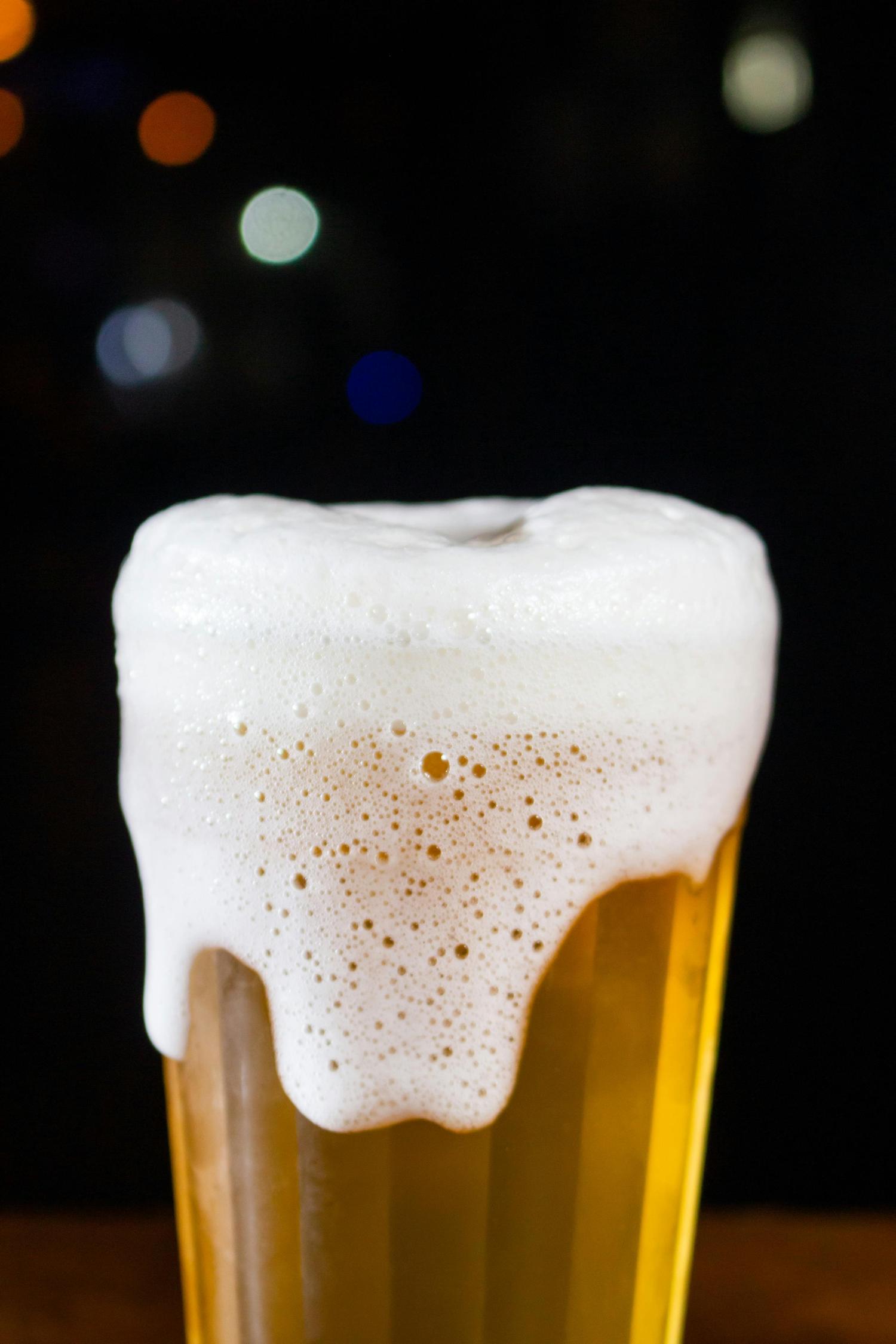 A glass of golden beer features a thick, frothy white head that overflows down the sides. The background is dark and blurred with scattered colorful bokeh lights.