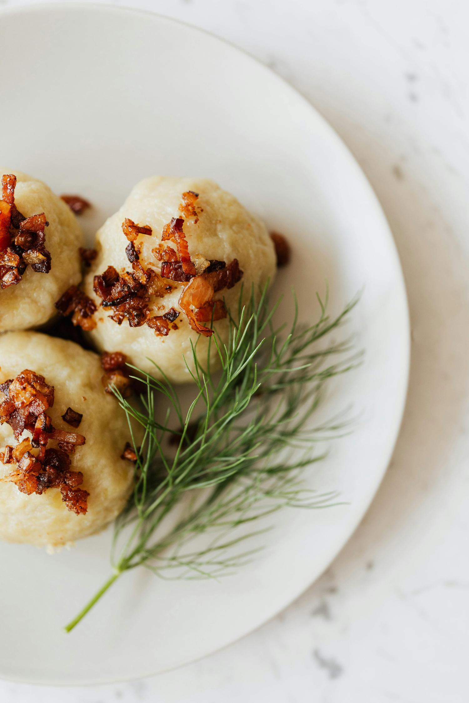 Potato dumplings topped with crispy fried bacon bits and a sprig of fresh dill are served on a white plate.