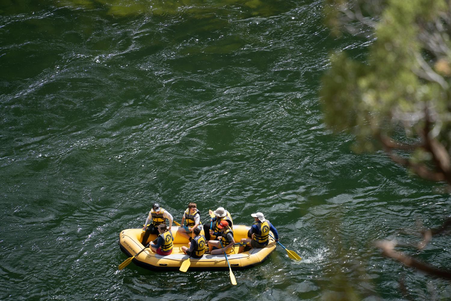 Six people in a yellow inflatable raft, wearing life vests and holding paddles, navigate deep green water.