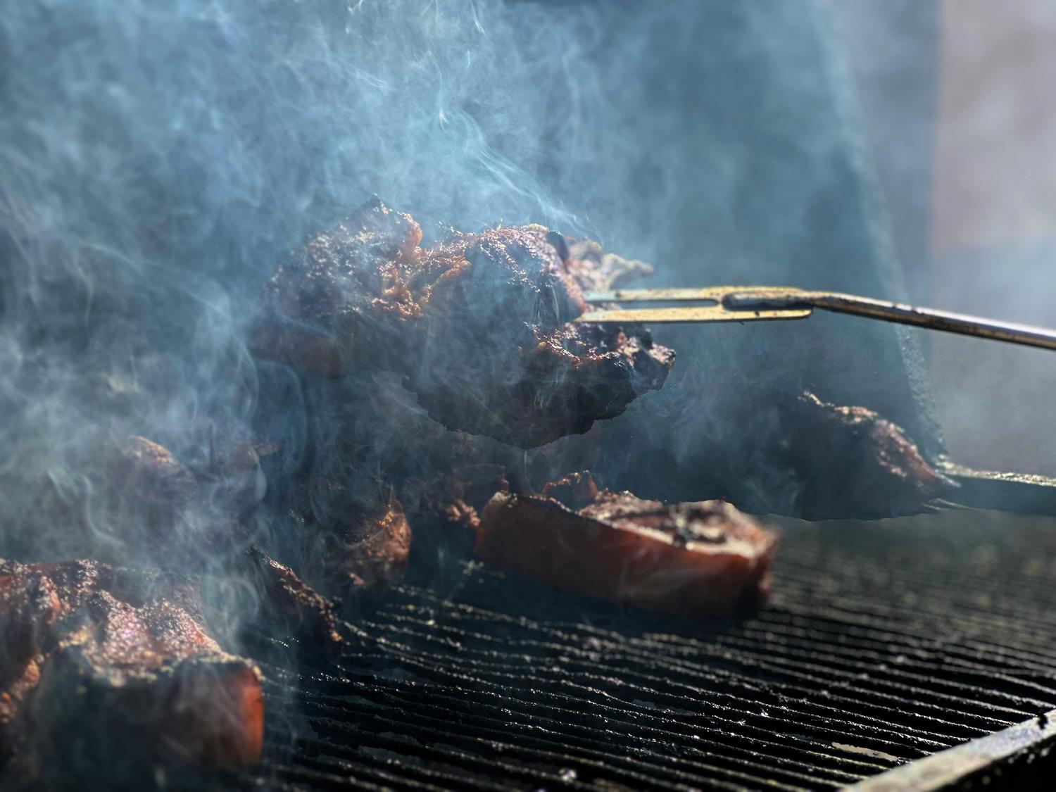 Seasoned cuts of meat sizzle and smoke heavily on a grill, as a pair of tongs adjusts one piece.