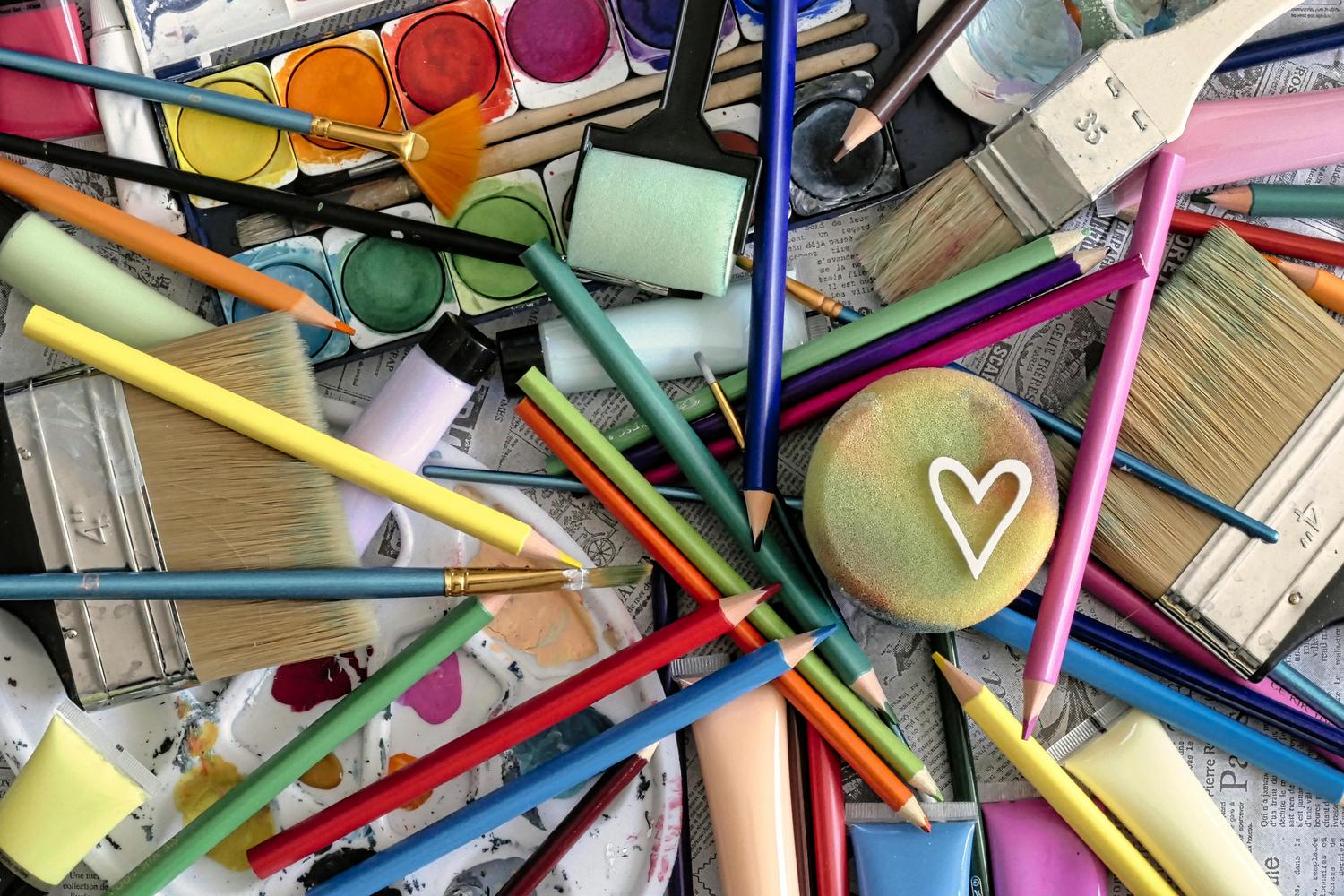 A vibrant collection of art supplies, including watercolor paints, brushes, colored pencils, and paint tubes, scattered across a surface, with one sponge featuring a white heart outline.