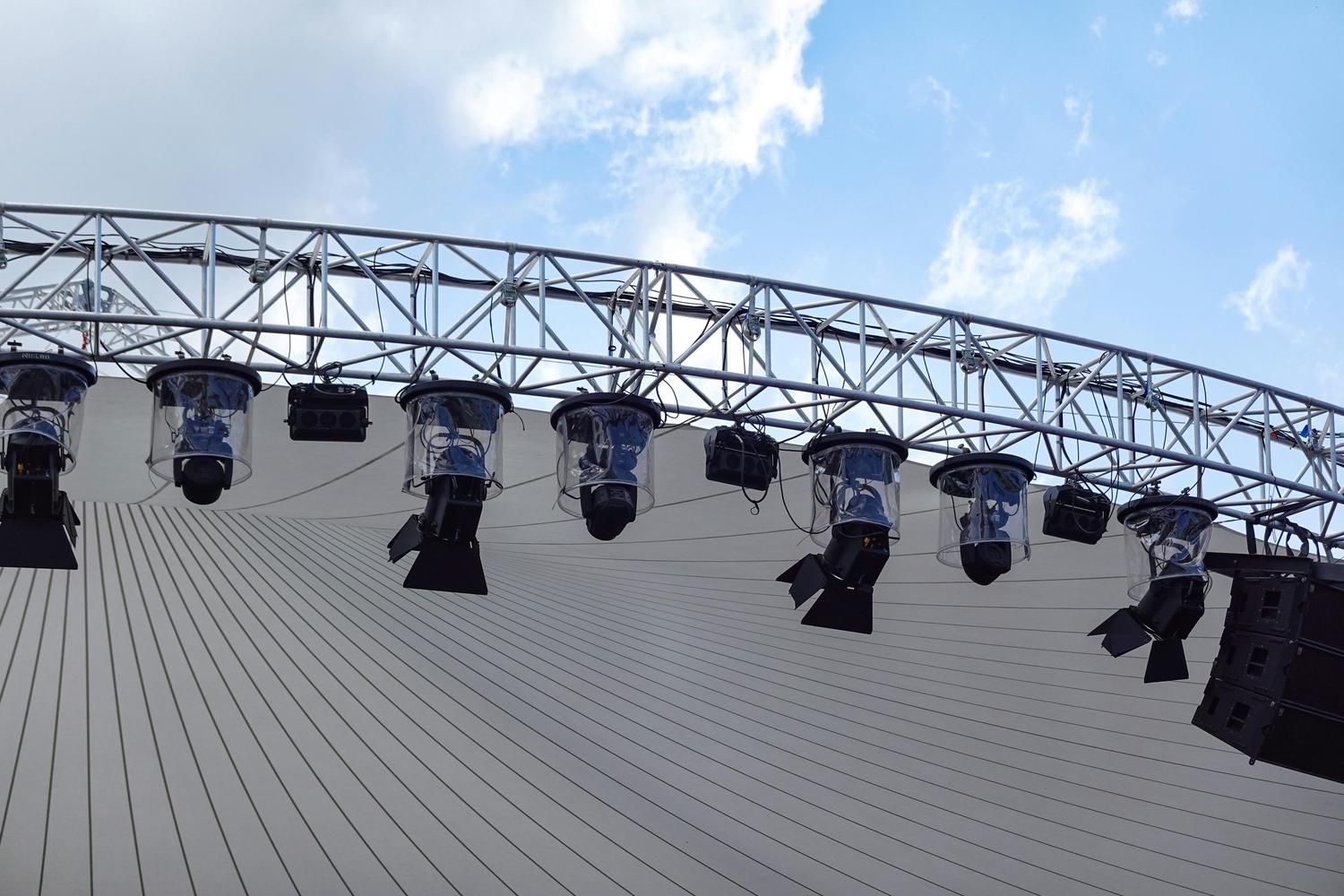 An arched metal truss supports professional stage lights, some with clear weather covers, and dark speaker arrays above a textured white canopy. A blue sky with light clouds is visible in the upper background, indicating an outdoor event setup.