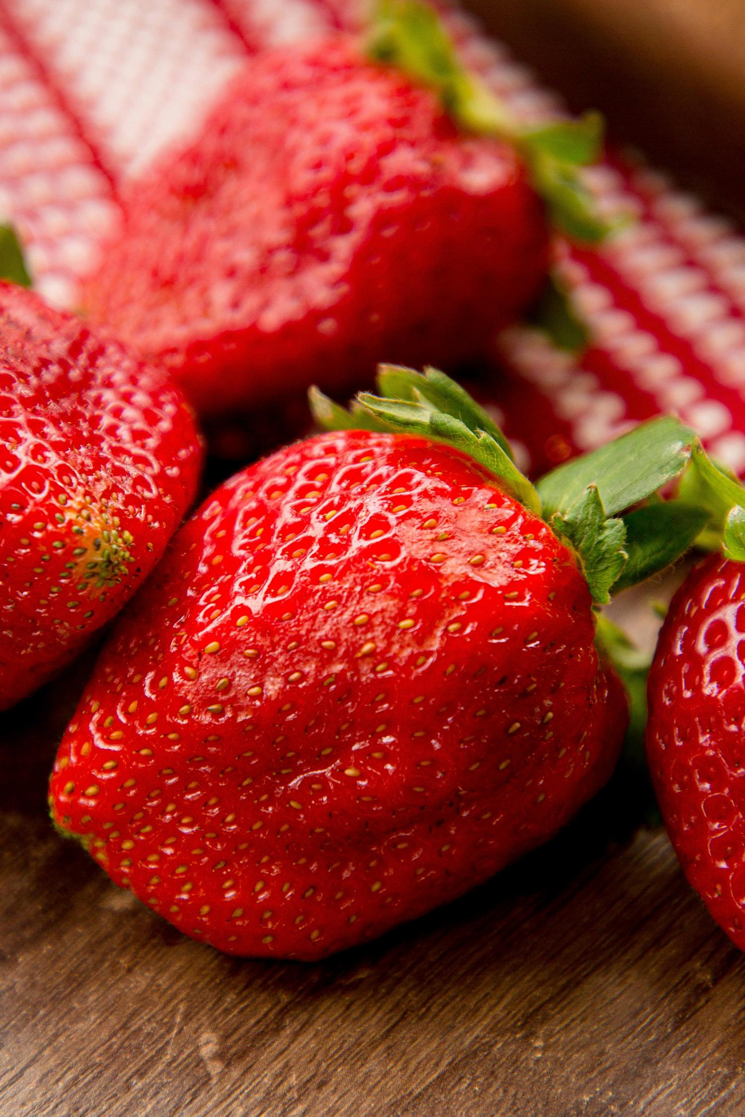 Close-up of several bright red strawberries with green leafy caps, resting on a rustic wooden surface with a blurred red and white checkered fabric in the background.