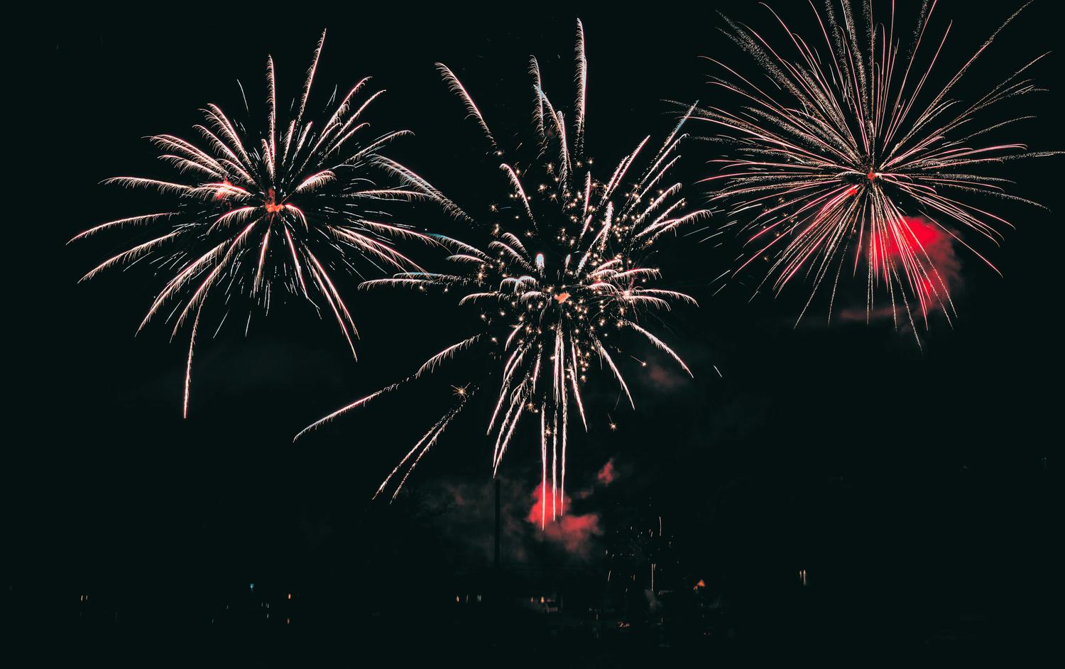 Three bright fireworks explode in a dark night sky, radiating white and gold streaks with some red illumination and smoke near their bases.