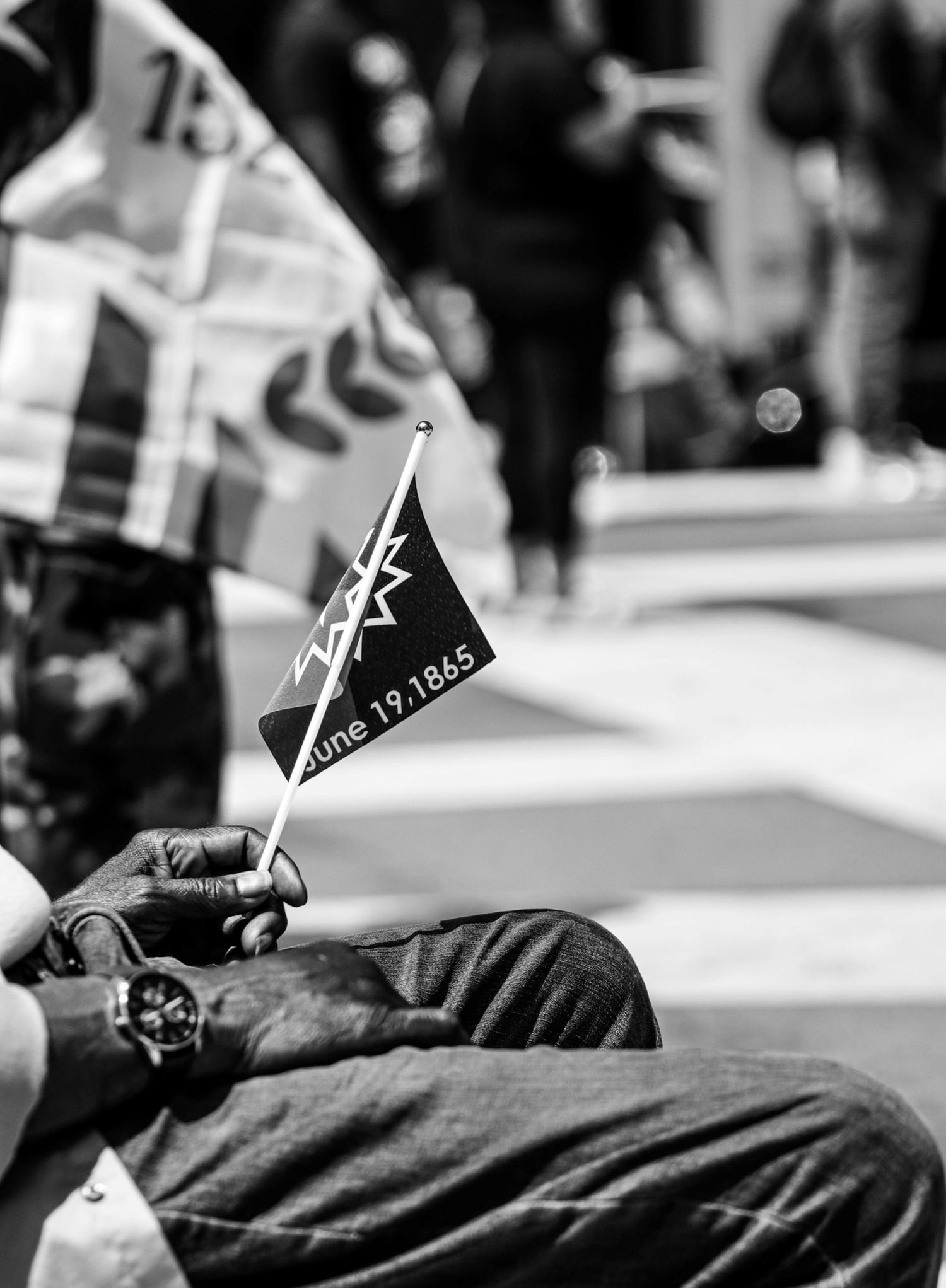 A person holds a small black Juneteenth flag featuring a white jagged design and the date 
