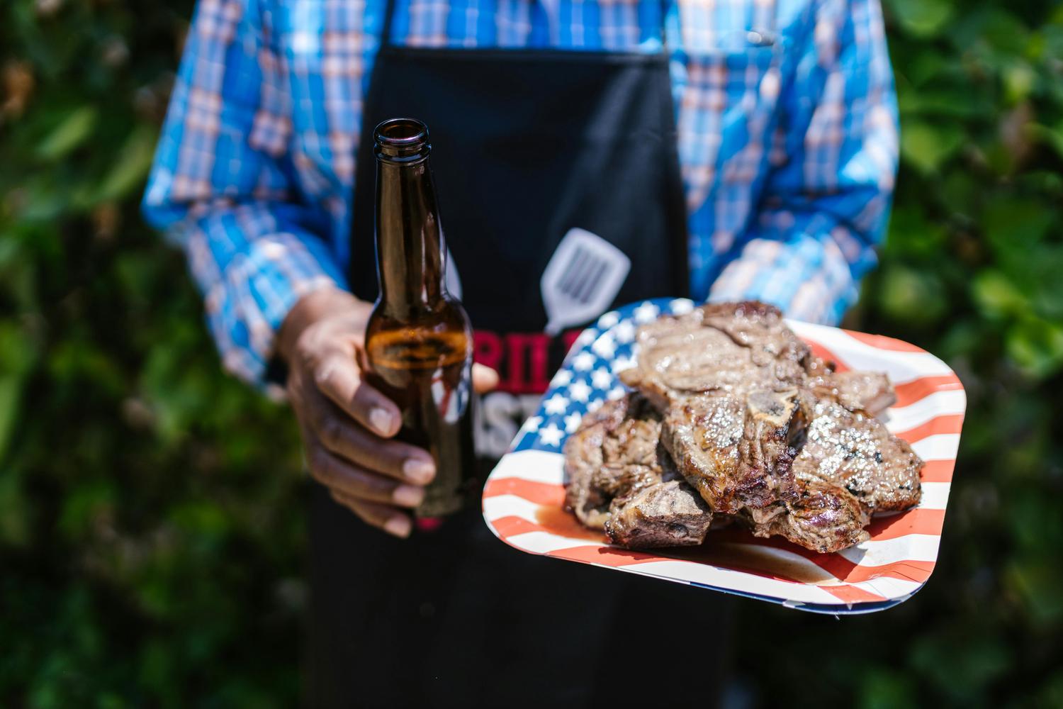 A person in a blue plaid shirt and black apron holds a brown beer bottle and a plate piled with grilled meat. The plate features a patriotic design with red and white stripes and blue stars.