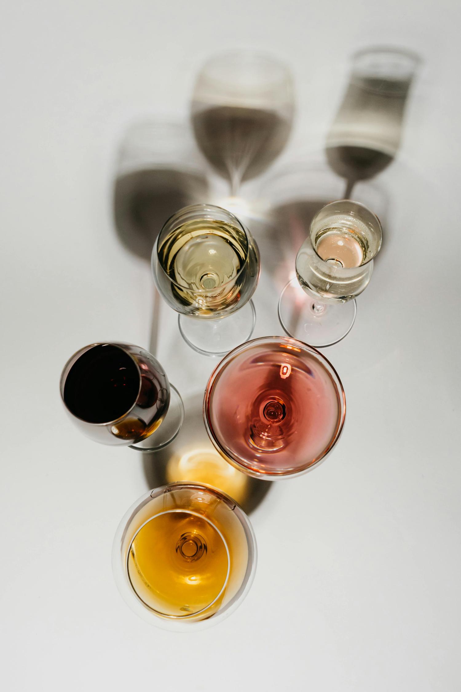 An overhead view of five wine glasses filled with white, rosé, red, and amber wines, casting long shadows on a white surface.