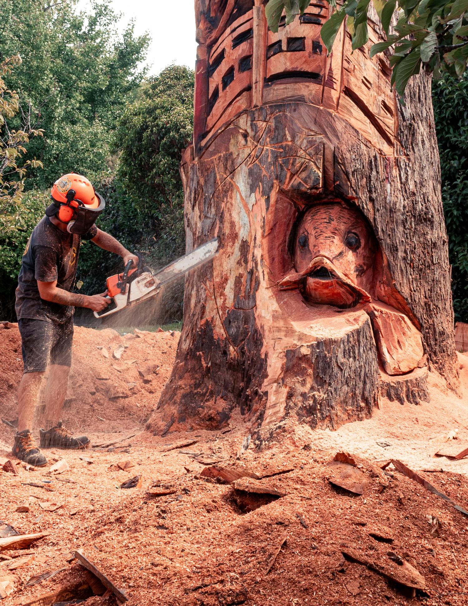 A wood carver in protective gear uses a chainsaw to sculpt a large tree trunk. The partially carved trunk reveals an intricate fish head design and other patterns, surrounded by piles of fresh sawdust.