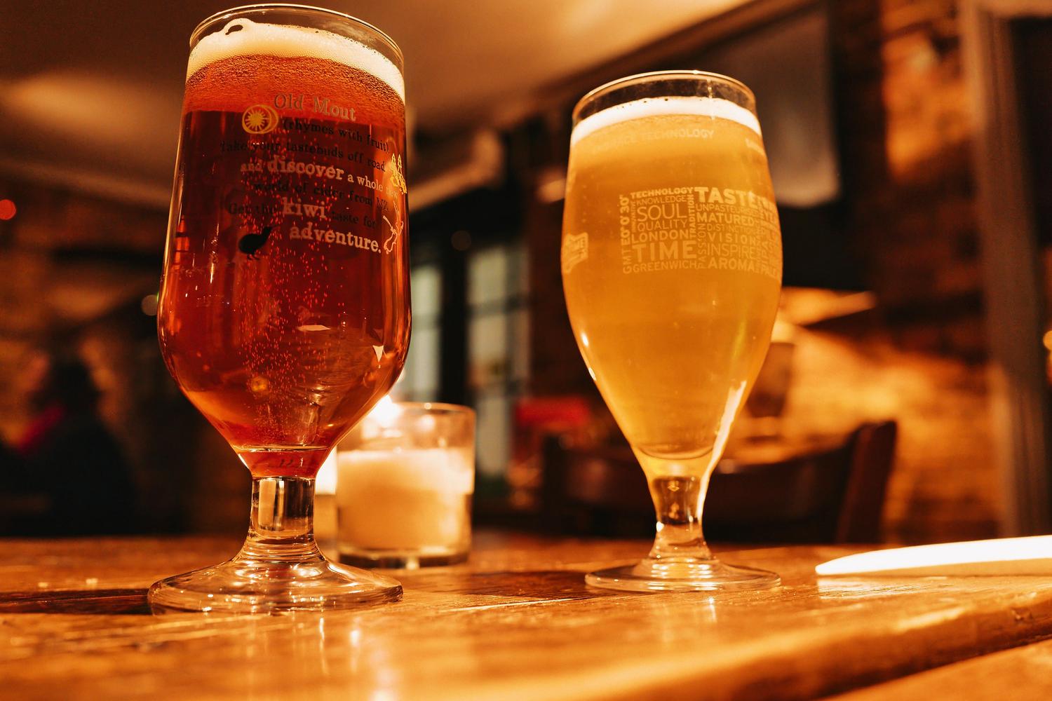 An amber Old Mout cider and a golden beer, both with foamy heads, sit in tall glasses on a warm wooden table in a dimly lit pub.
