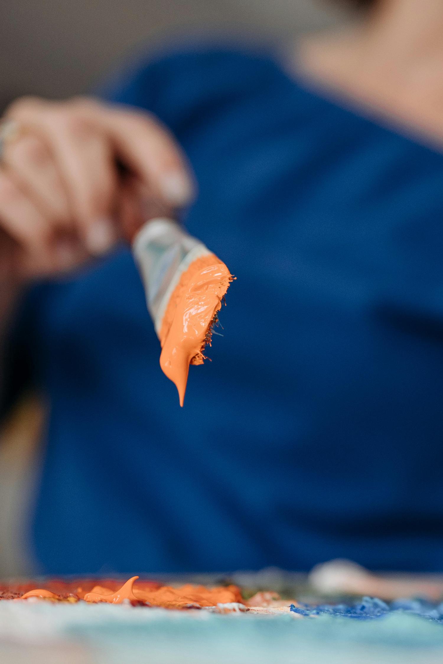 A close-up shows an artist's brush loaded with vibrant orange paint, a drip forming at its tip. A blurred hand holds the brush above a paint palette, with a blue background.