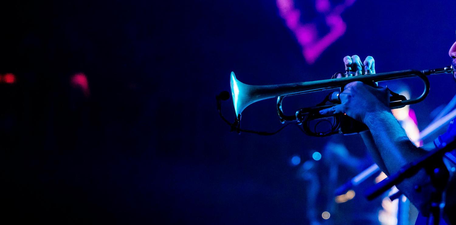 A musician's hands play a trumpet, brightly illuminated by blue stage lights against a dark, blurry background with hints of purple and blue.