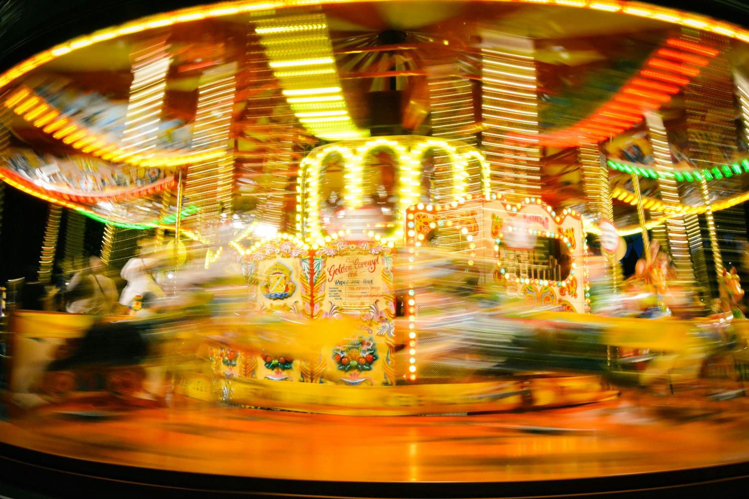 A brightly lit carousel spins rapidly at night, creating streaks of vibrant yellow, red, and green light from the motion blur. Blurred figures are visible on the ride, conveying a dynamic, festive atmosphere.
