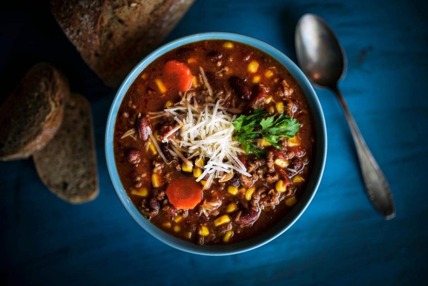 A bowl of hearty chili or stew, featuring ground meat, beans, corn, and carrots, is topped with shredded cheese and a sprig of parsley. It sits on a dark blue surface next to slices of rustic bread and a silver spoon.