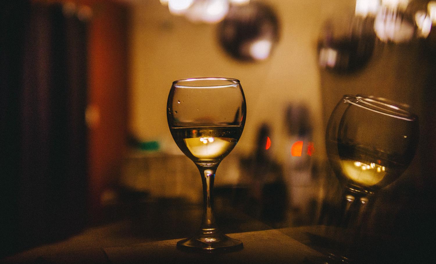 A clear wine glass filled with light-colored liquid stands centered, reflecting warm lights from a blurry background. Another out-of-focus glass is visible to the right.