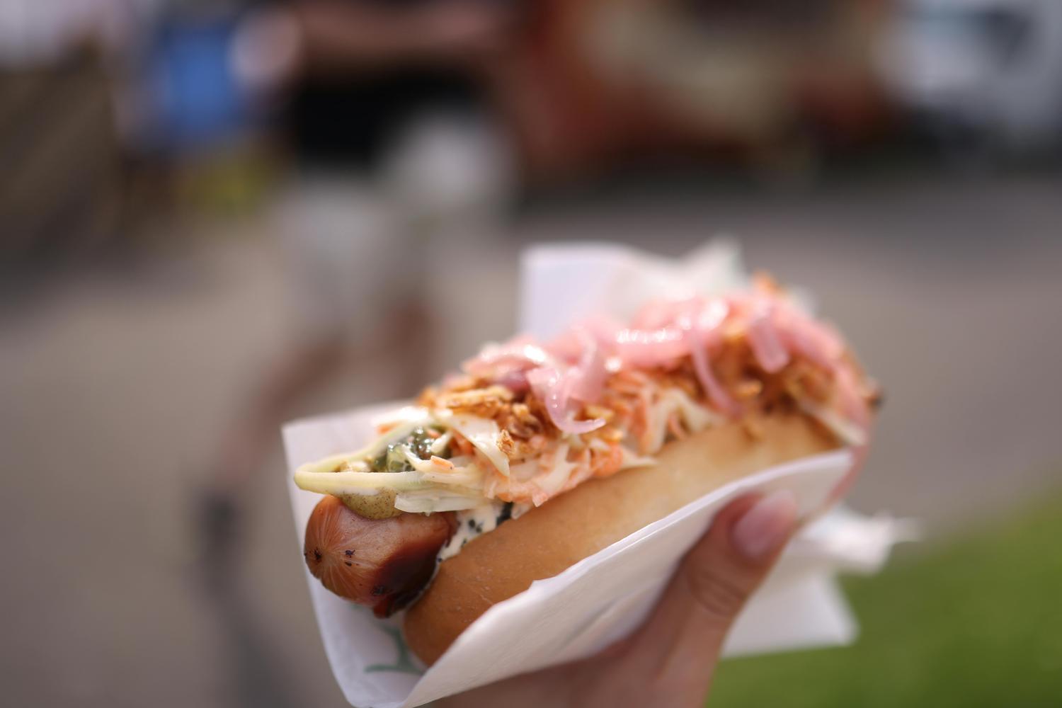 A hand holds a hot dog loaded with a sausage, light green sauce, white coleslaw, crispy fried onions, and pink pickled onions in a bun. The background is softly blurred, suggesting an outdoor food event.
