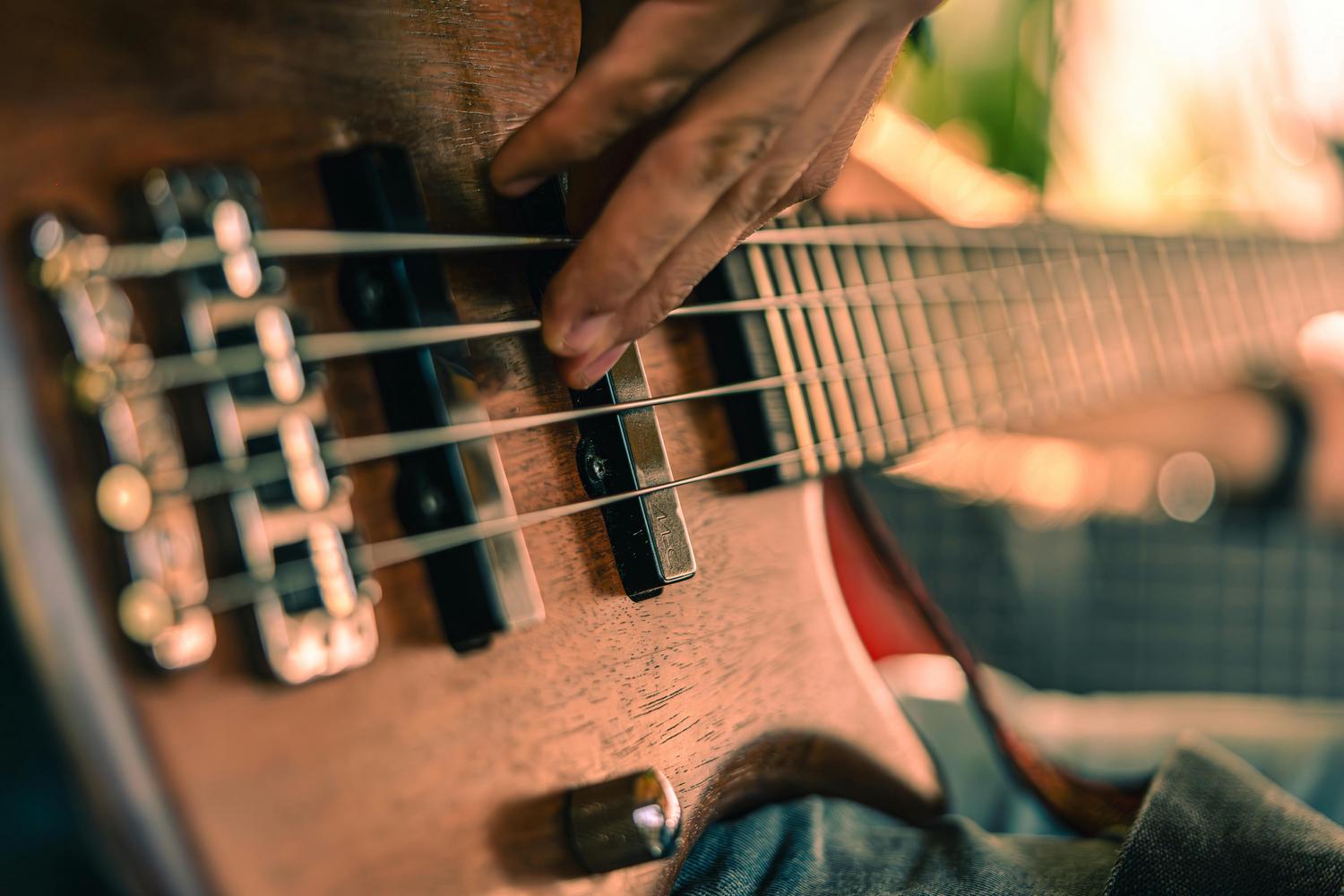 A close-up shows a hand plucking the strings of a wooden electric bass guitar, with the fretboard and a hint of blue jeans blurred in the background.