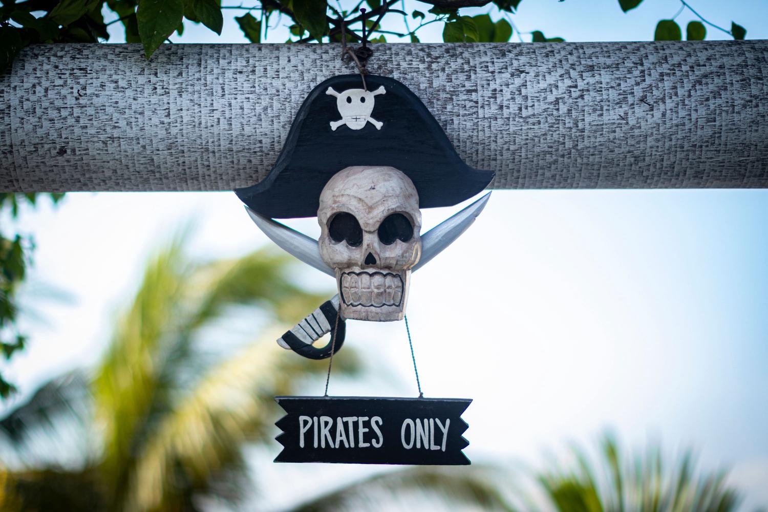 A whimsical wooden pirate skull wearing a black hat with a skull and crossbones emblem hangs with crossed cutlasses and a sign that reads 