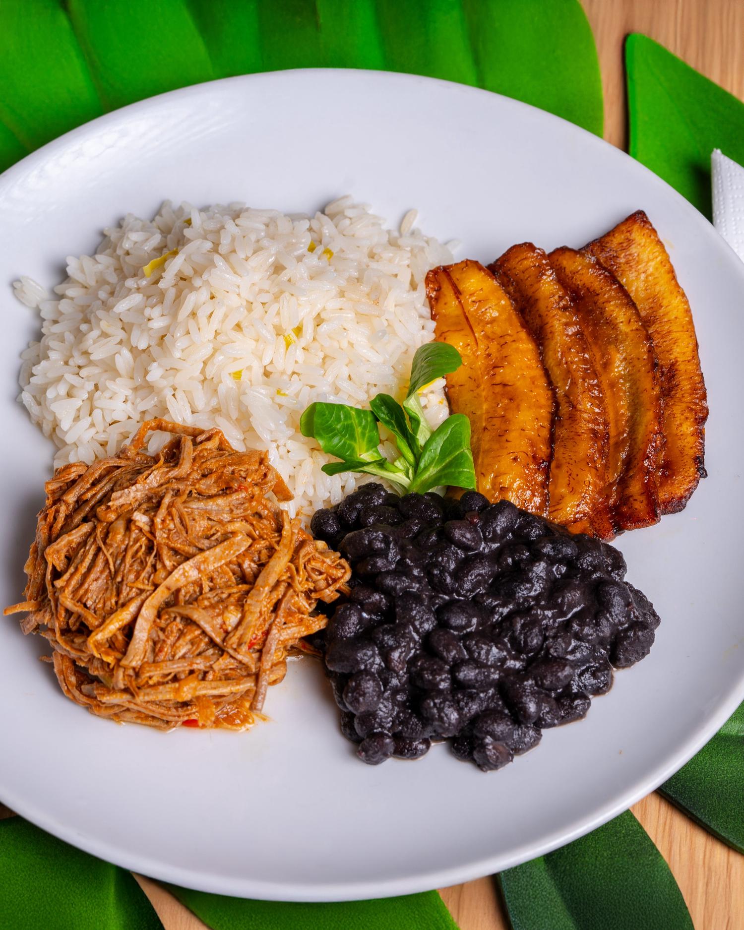 A white plate displays a meal of fluffy white rice, saucy shredded meat, black beans, and glistening fried plantains, garnished with a small green sprig.