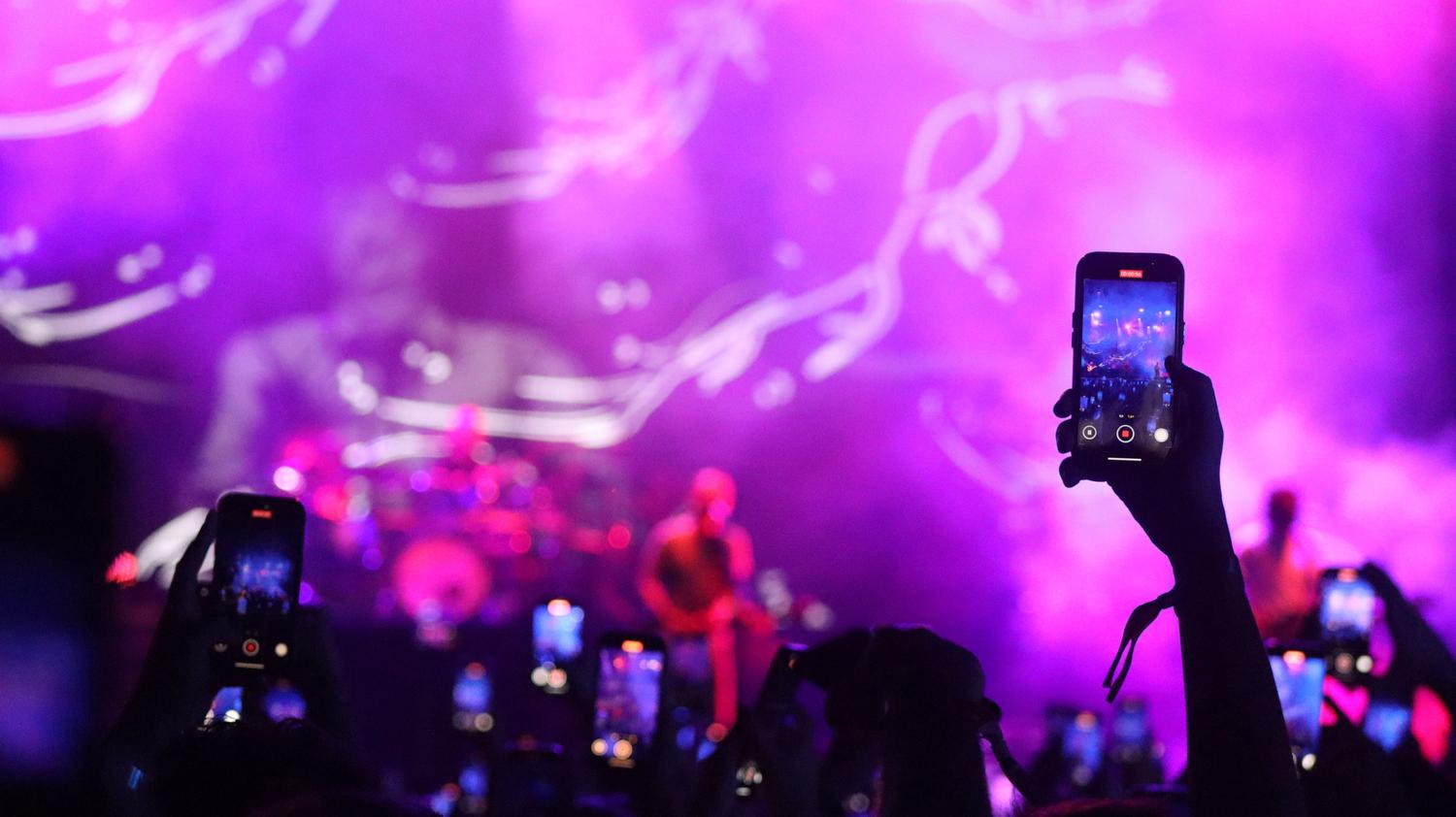 A vibrant purple and pink-lit concert scene shows a crowd holding up smartphones to record, with a prominent hand in the foreground actively capturing the performance.