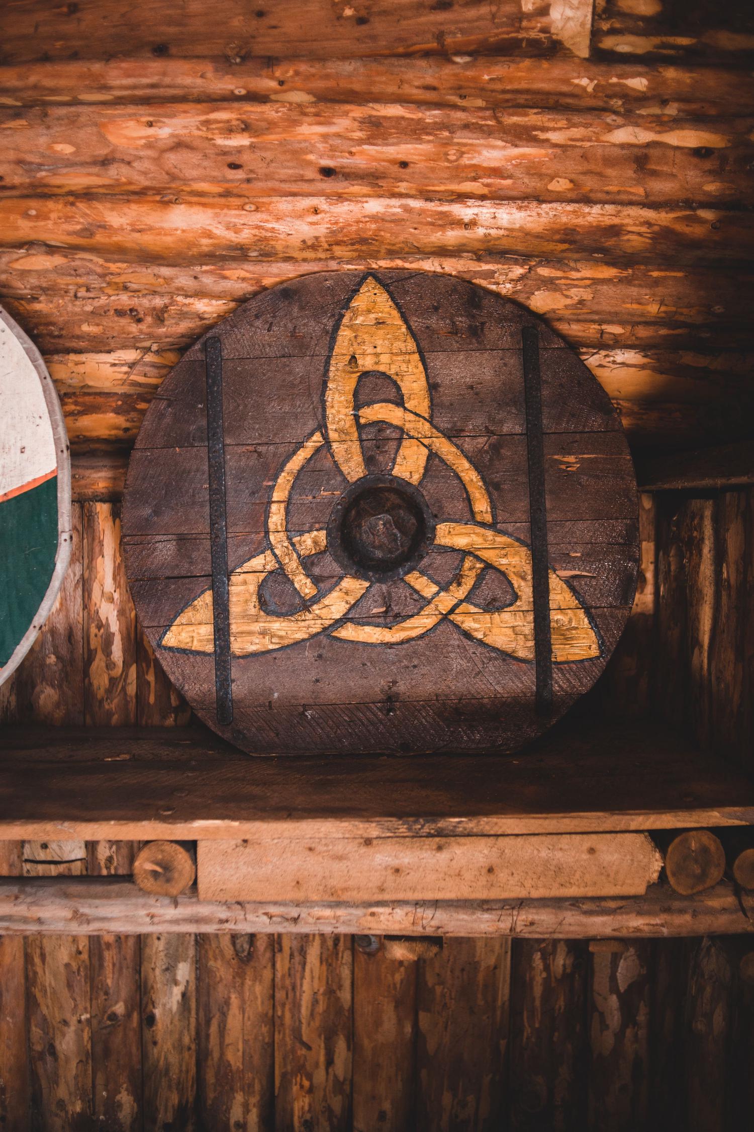 A round, dark wooden shield with a gold Celtic knot design and a prominent metal boss rests on a rustic wooden shelf. A log wall forms the background, with part of another shield visible on the left.
