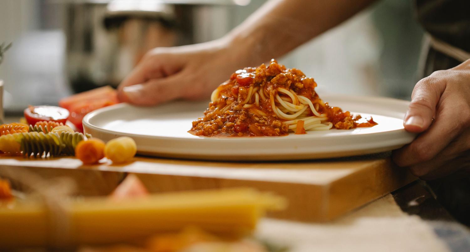 A person's hands gently hold a white plate of spaghetti topped with a rich red meat sauce. Blurred in the foreground are various uncooked pasta shapes and fresh tomatoes, suggesting a kitchen setting.