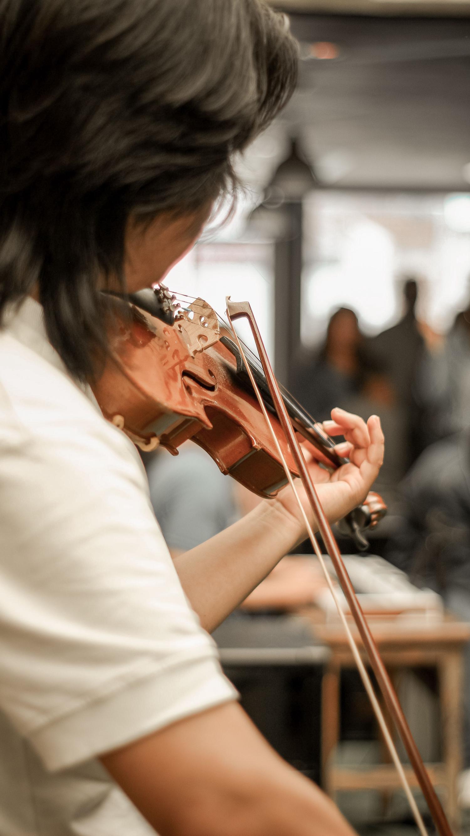 A person with dark hair plays a violin, with a bow drawn across the strings. Blurred figures and a cafe setting are visible in the soft-focus background.