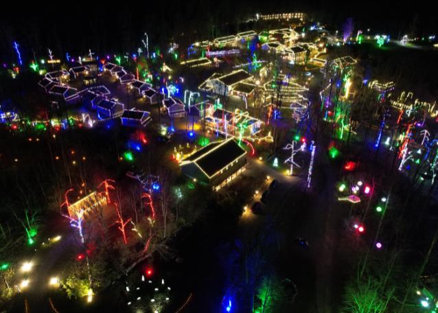 An aerial nighttime view of a sprawling outdoor holiday light display, featuring numerous buildings and trees illuminated with bright, multi-colored lights.