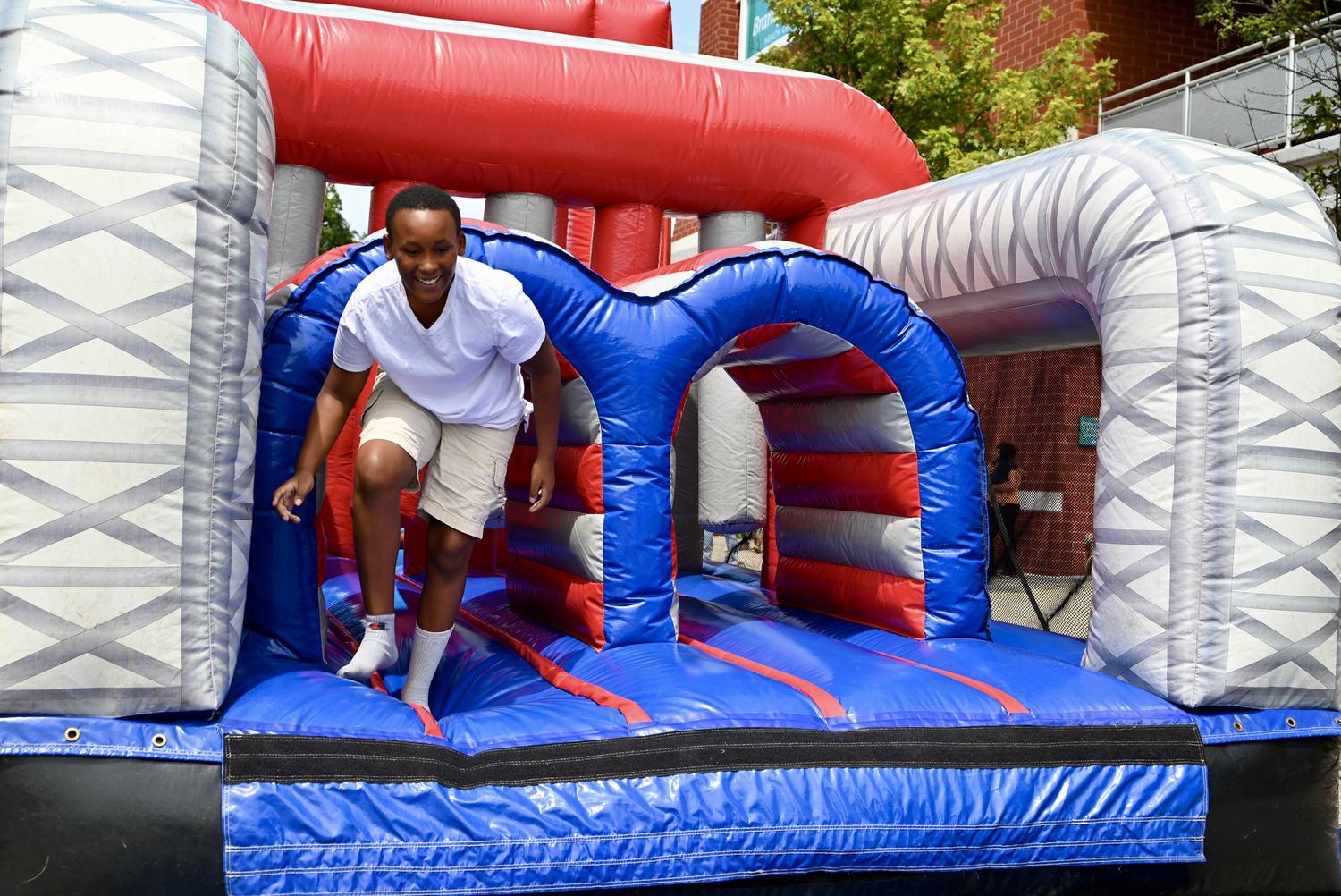 A smiling boy in a white t-shirt and khaki shorts steps through a blue arch on a large red, white, and blue inflatable obstacle course. He appears to be having fun at an outdoor event.