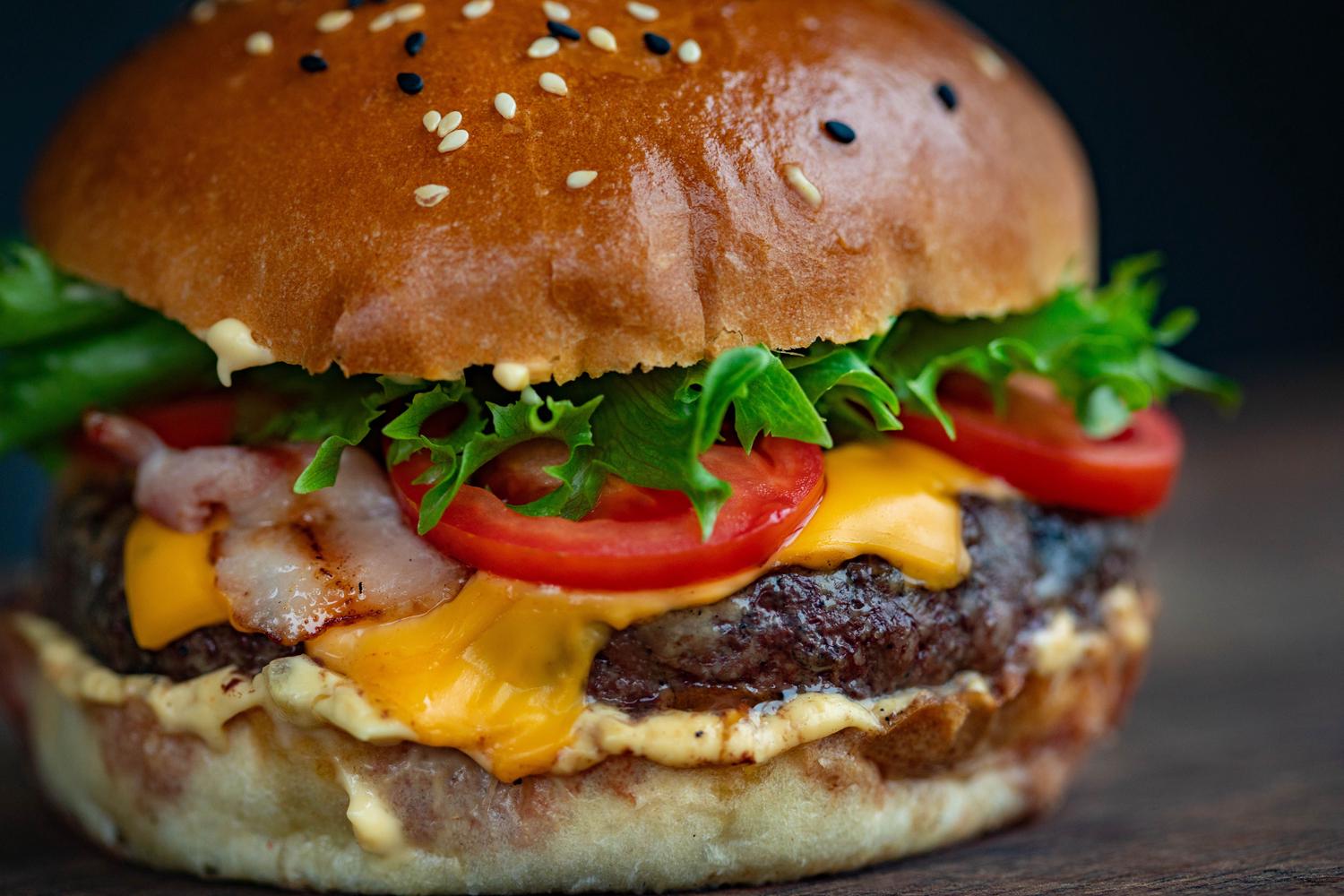 A close-up of a loaded burger with a sesame seed bun, beef patty, melted yellow cheese, crispy bacon, fresh lettuce, and red tomato slices.