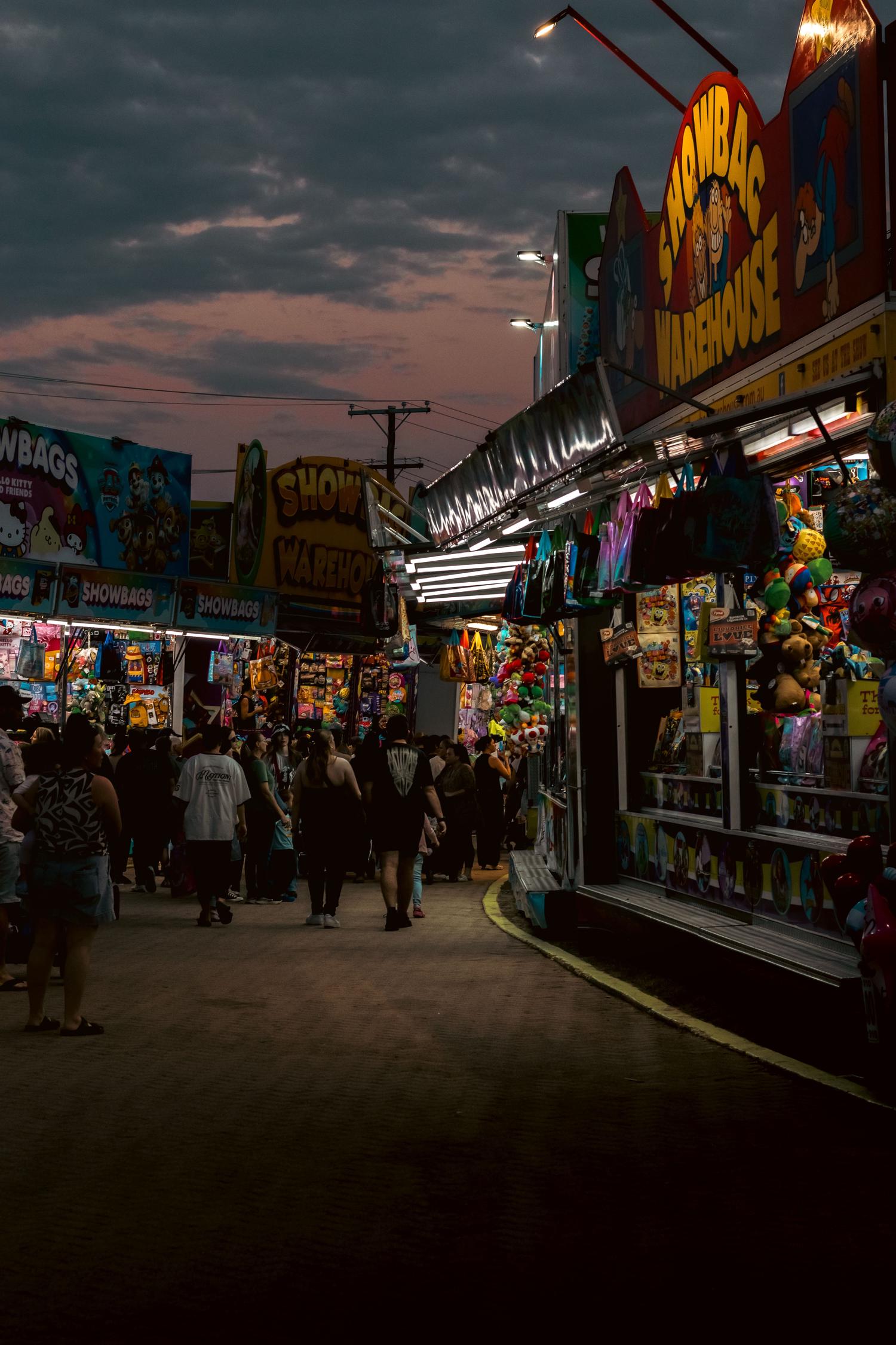 A bustling night market scene at dusk, with numerous people walking past brightly lit 