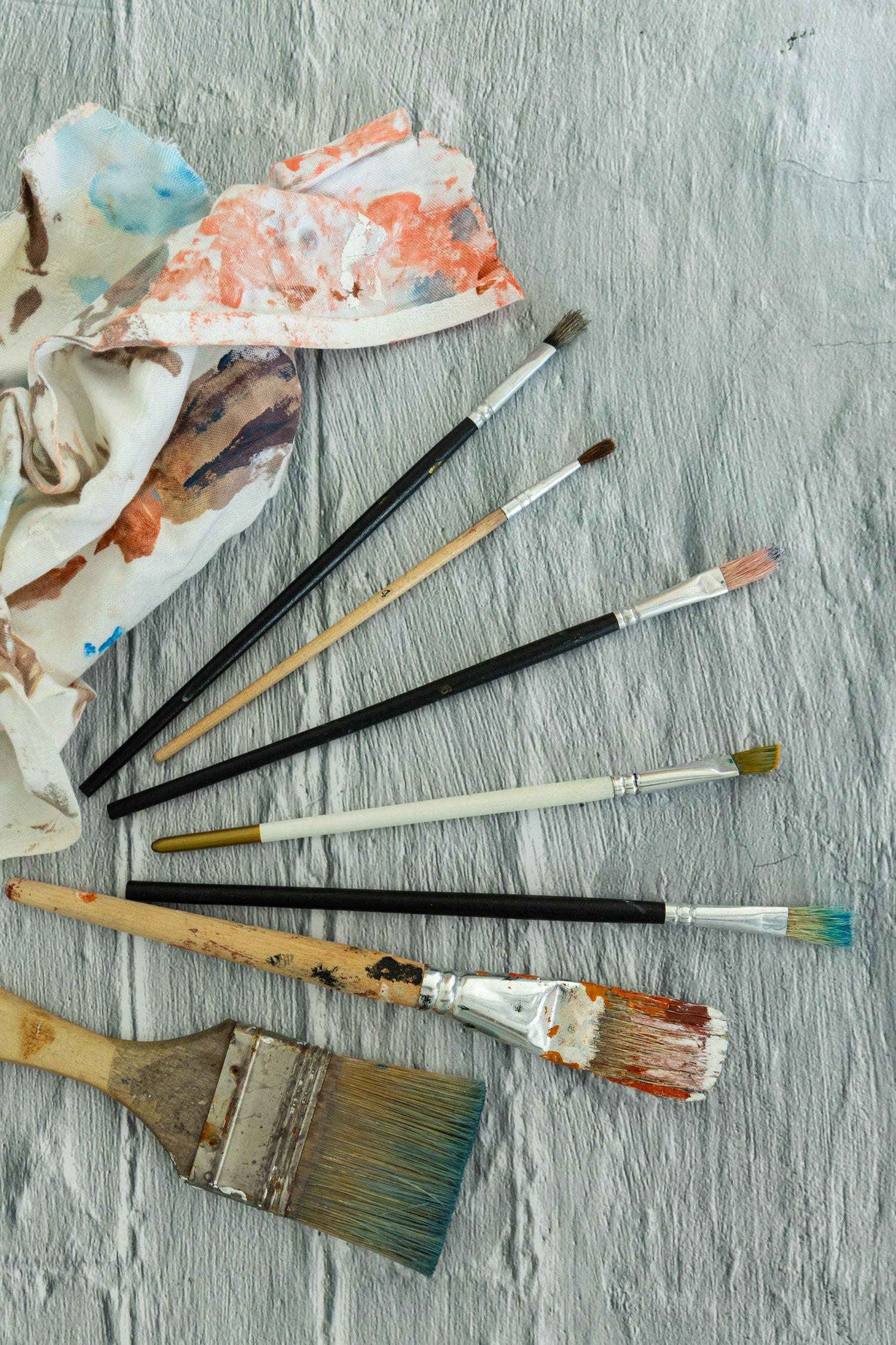 Various paintbrushes, some stained with different colored paints, are arranged on a textured grey surface next to a paint-splattered cloth.