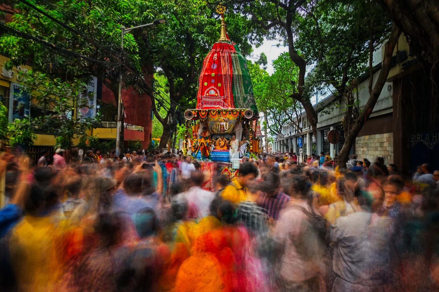 A vibrant street procession features a large, elaborately decorated red and green chariot, surrounded by a dense, blurry crowd of people moving amidst lush green trees and buildings.