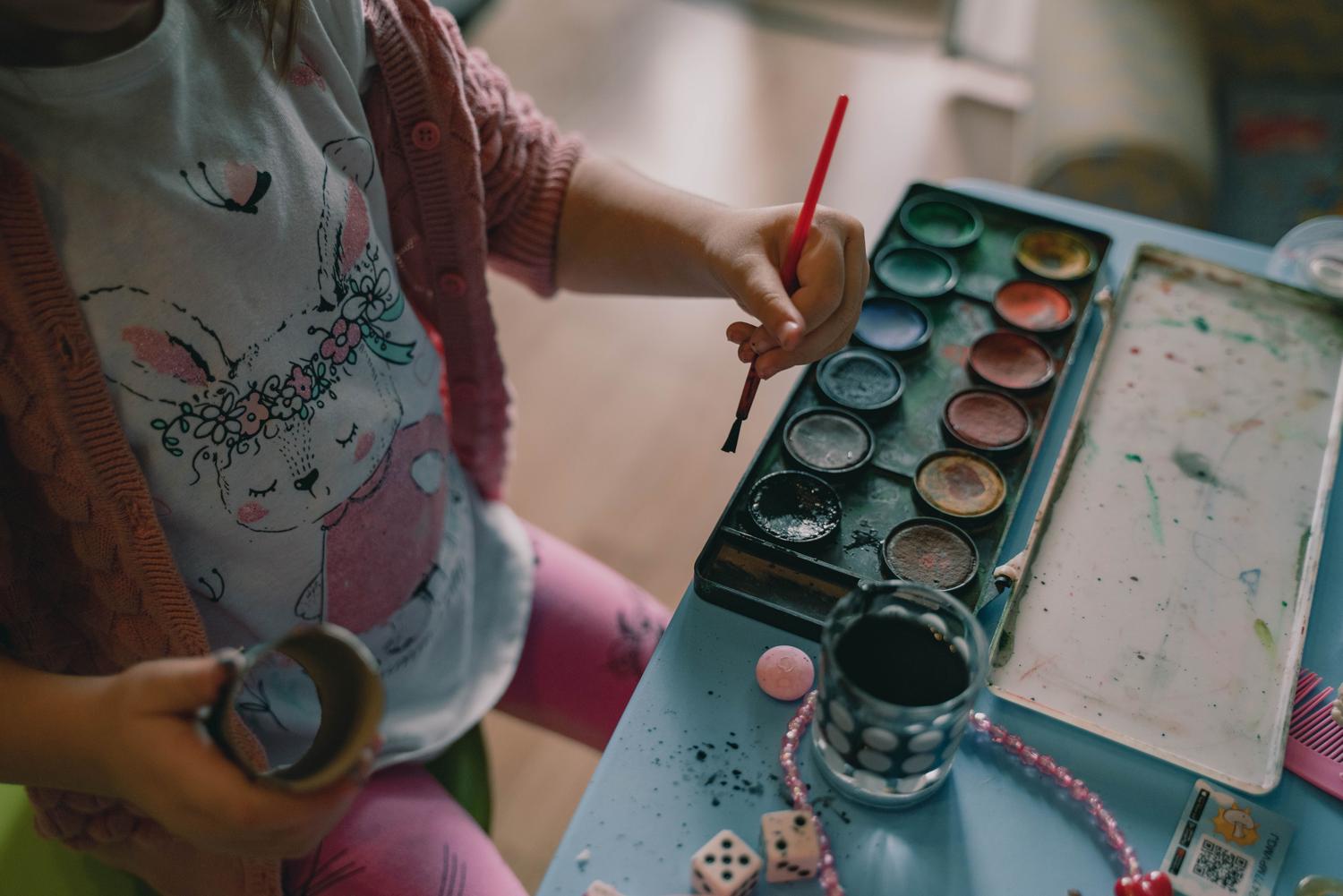 A child in a bunny-print shirt and pink cardigan holds a red paintbrush above a colorful watercolor palette, poised to paint on a messy blue table surrounded by art supplies and small toys.