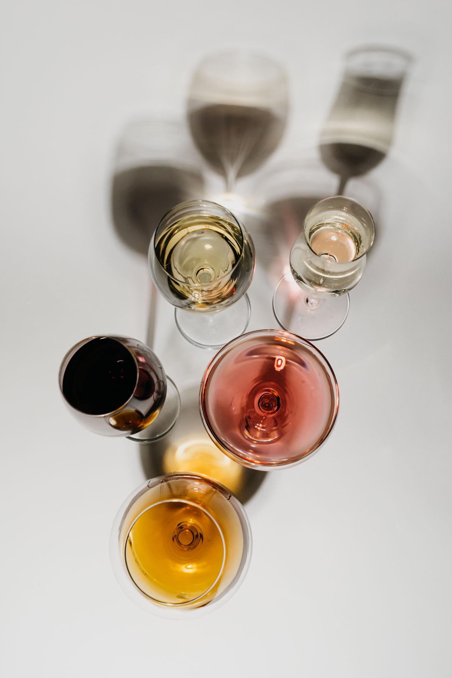 Five wine glasses, seen from above, hold white, rosé, red, and amber-colored wines. They cast prominent shadows on a bright white surface.