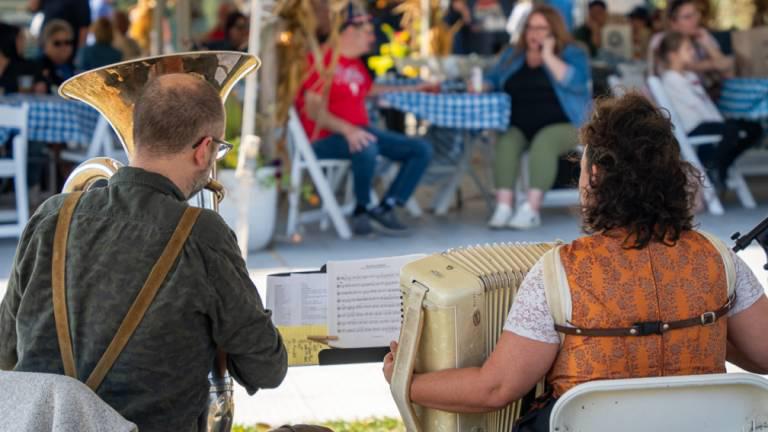 Two musicians, one playing a tuba and the other an accordion, are seen from behind performing at an outdoor festival where people sit at tables in the blurred background.