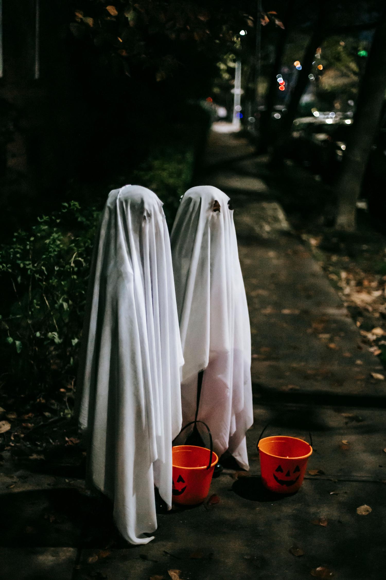 Two figures in white sheet ghost costumes with eyeholes stand on a dark path at night, each holding an orange pumpkin trick-or-treat bucket.