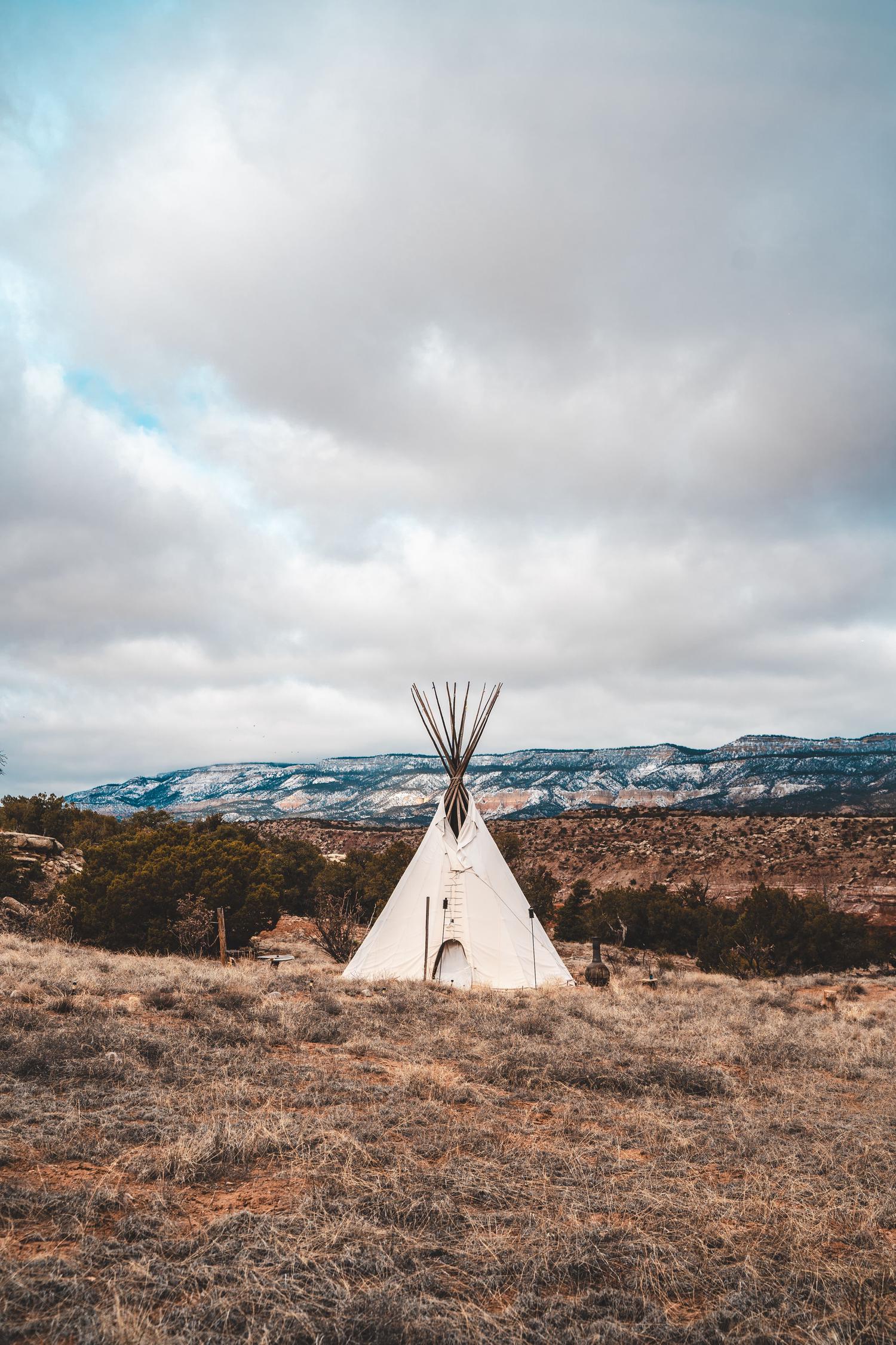 A white tipi with prominent wooden poles stands in a dry, grassy field. In the background are snow-dusted mountains and red rock formations under a cloudy sky.