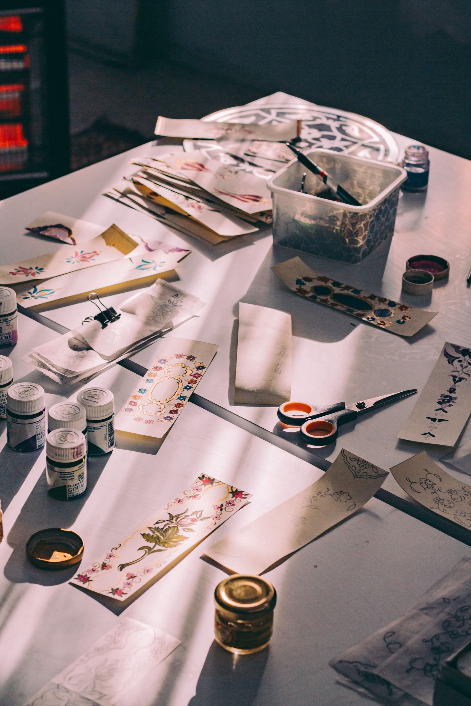 A sunlit white table covered with gouache paints, brushes, scissors, and small paper strips featuring intricate floral and ornamental designs in various stages of completion.