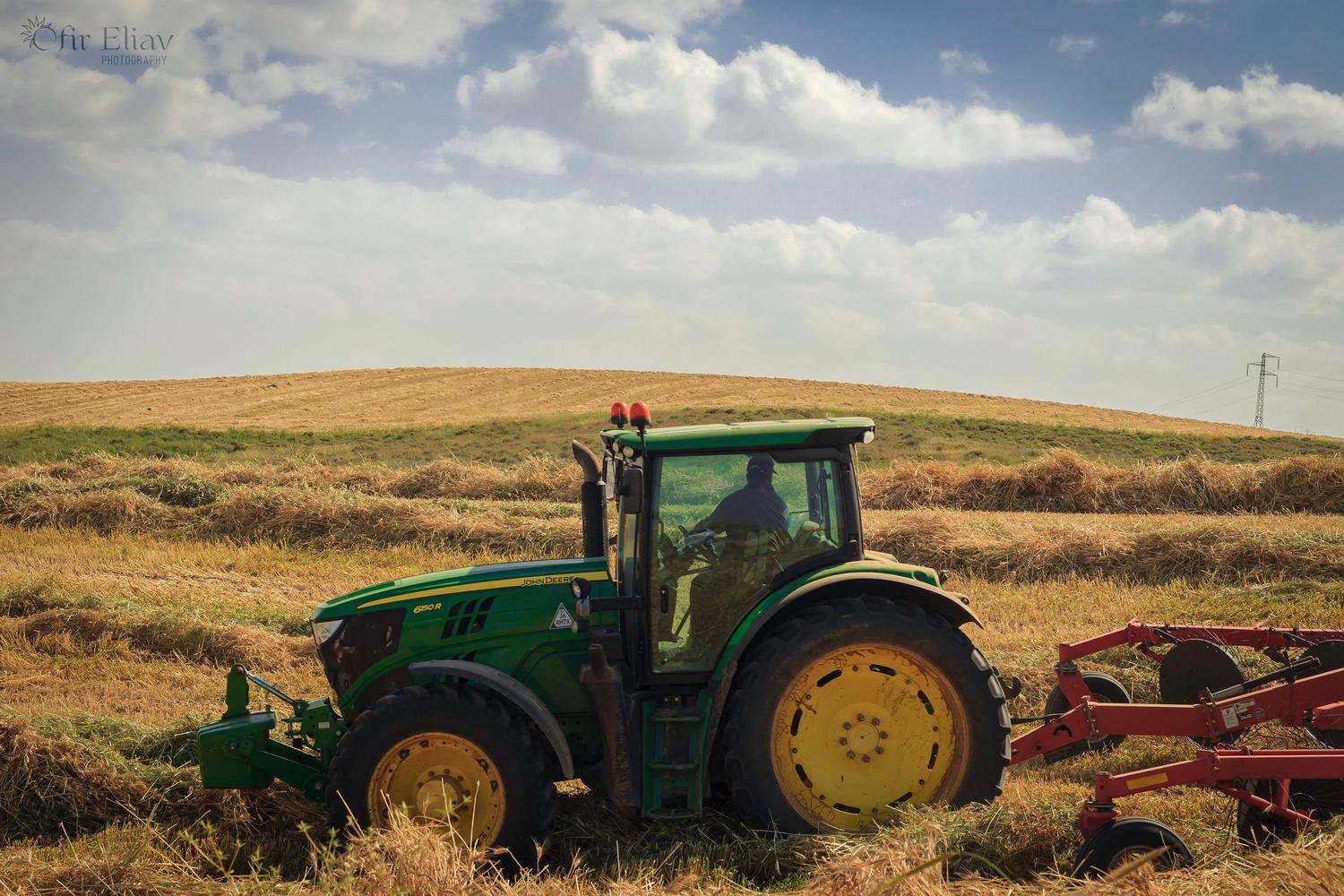 A green John Deere tractor with a visible driver works through a golden field of cut hay or straw, pulling a red farm implement under a partly cloudy sky.