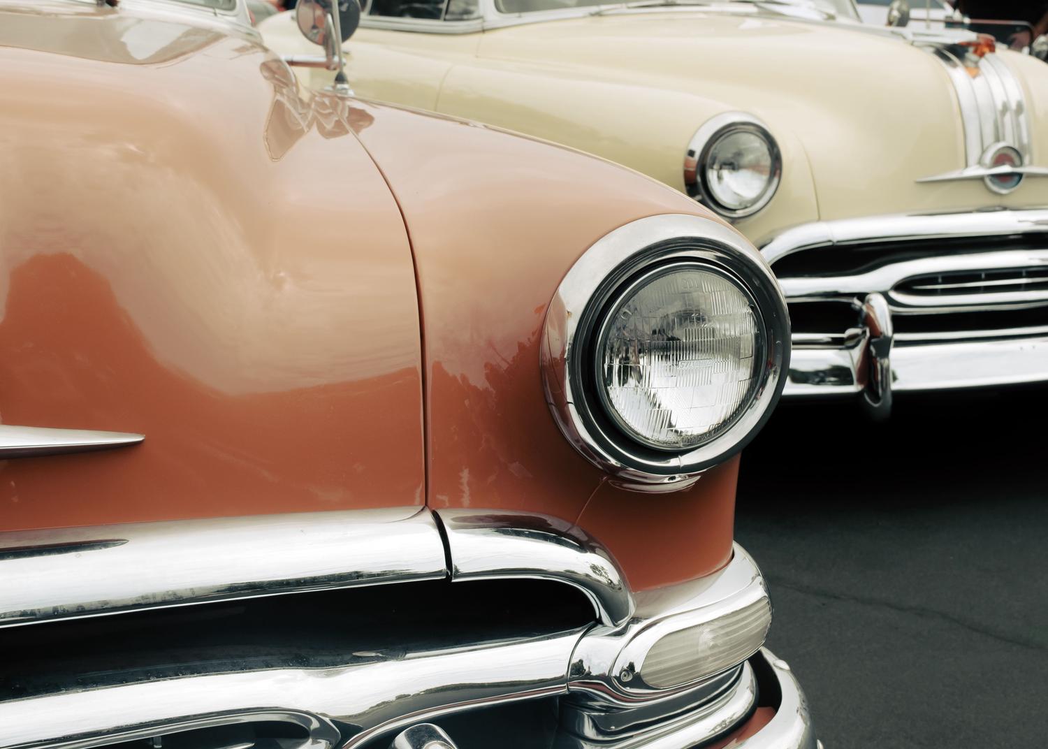 A close-up shows the front quarter panel of a peach vintage car with a round chrome headlight and bumper, with a cream-colored vintage car partially visible behind it.