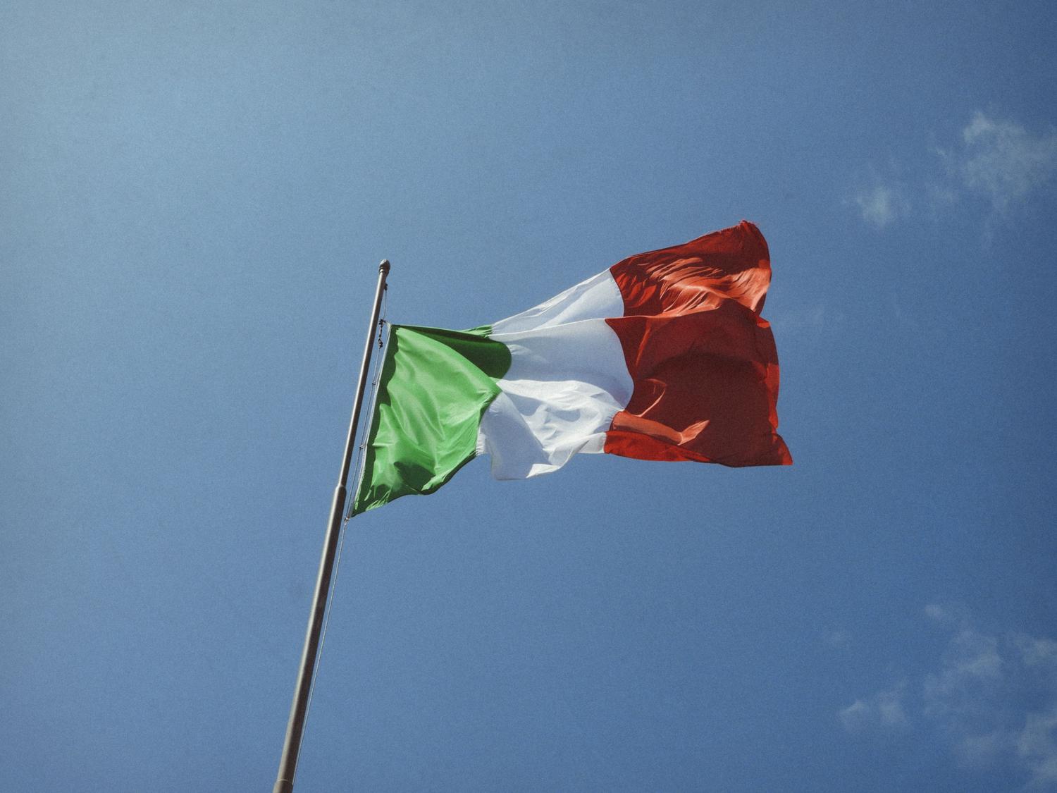 The Italian flag, with vertical stripes of green, white, and red, waves from a flagpole against a clear blue sky.