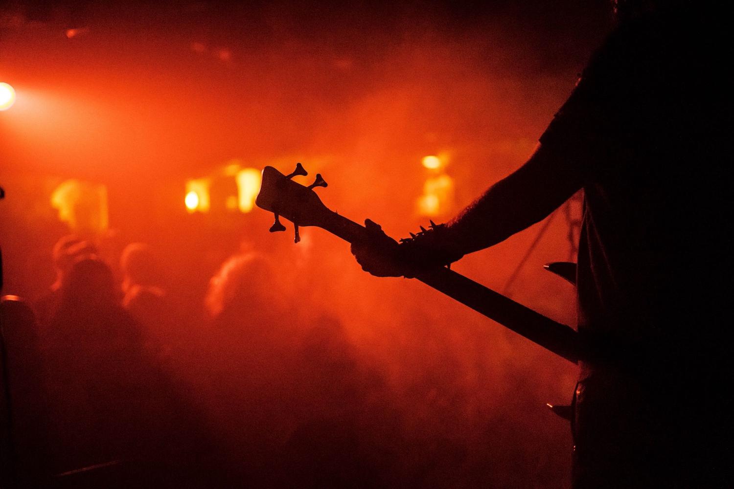 A musician's arm, adorned with spikes, holds a bass guitar, silhouetted against a vibrant, hazy orange-red stage light. Faint audience silhouettes are visible in the background, suggesting a live concert.