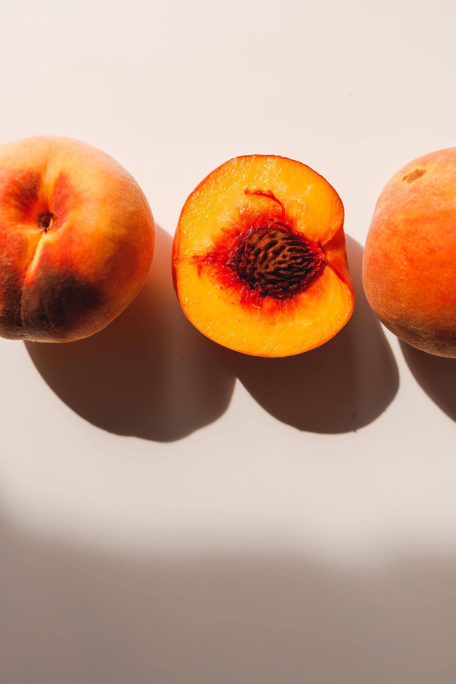 Two whole peaches flank a halved peach on a light surface. The central peach reveals its vibrant orange flesh and dark, ridged pit.