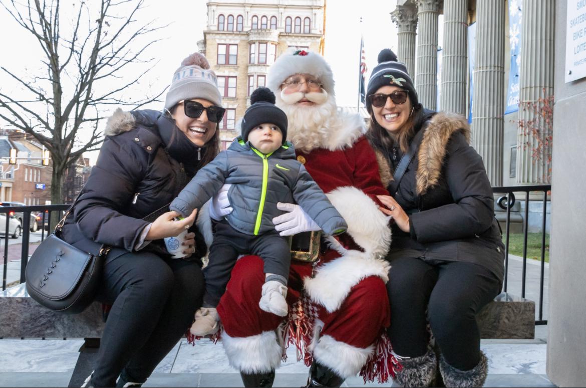 Santa Claus sits outdoors with a toddler on his lap, flanked by two smiling women wearing winter coats and hats, against a backdrop of a building.