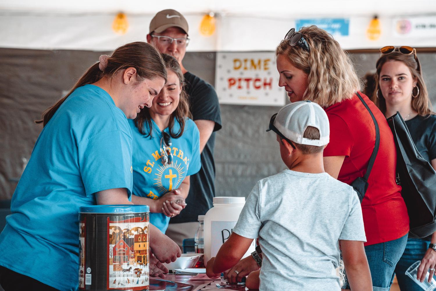 A group of adults and a child are gathered around a table under a tent, appearing to participate in an activity. A blurred sign in the background reads 