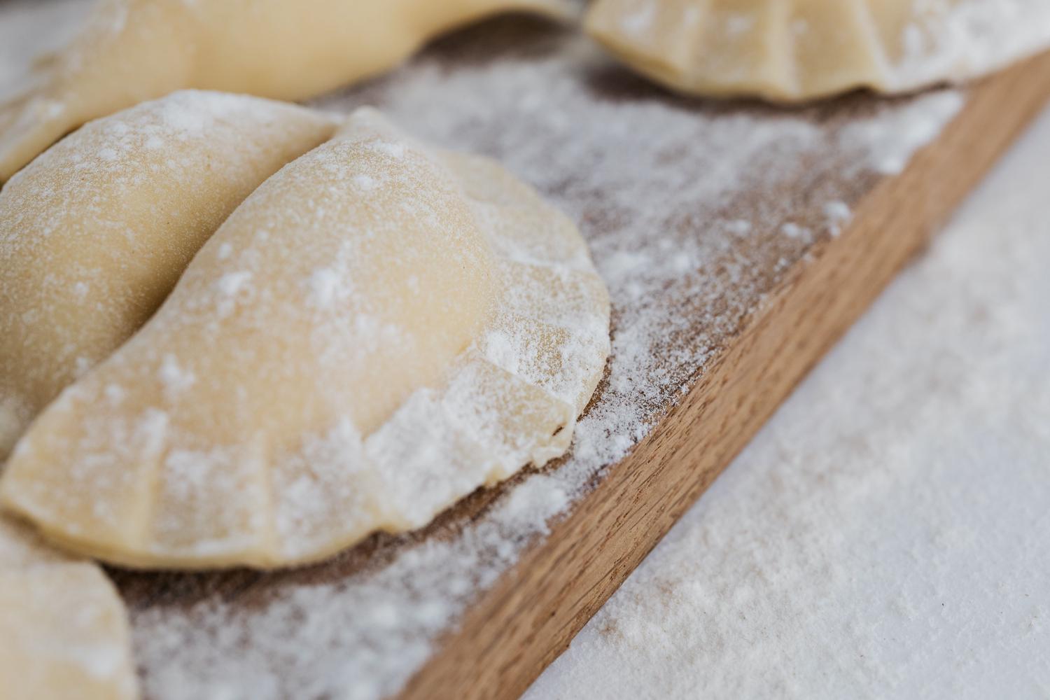 Several uncooked, crescent-shaped dumplings with crimped edges are dusted with flour on a wooden cutting board.
