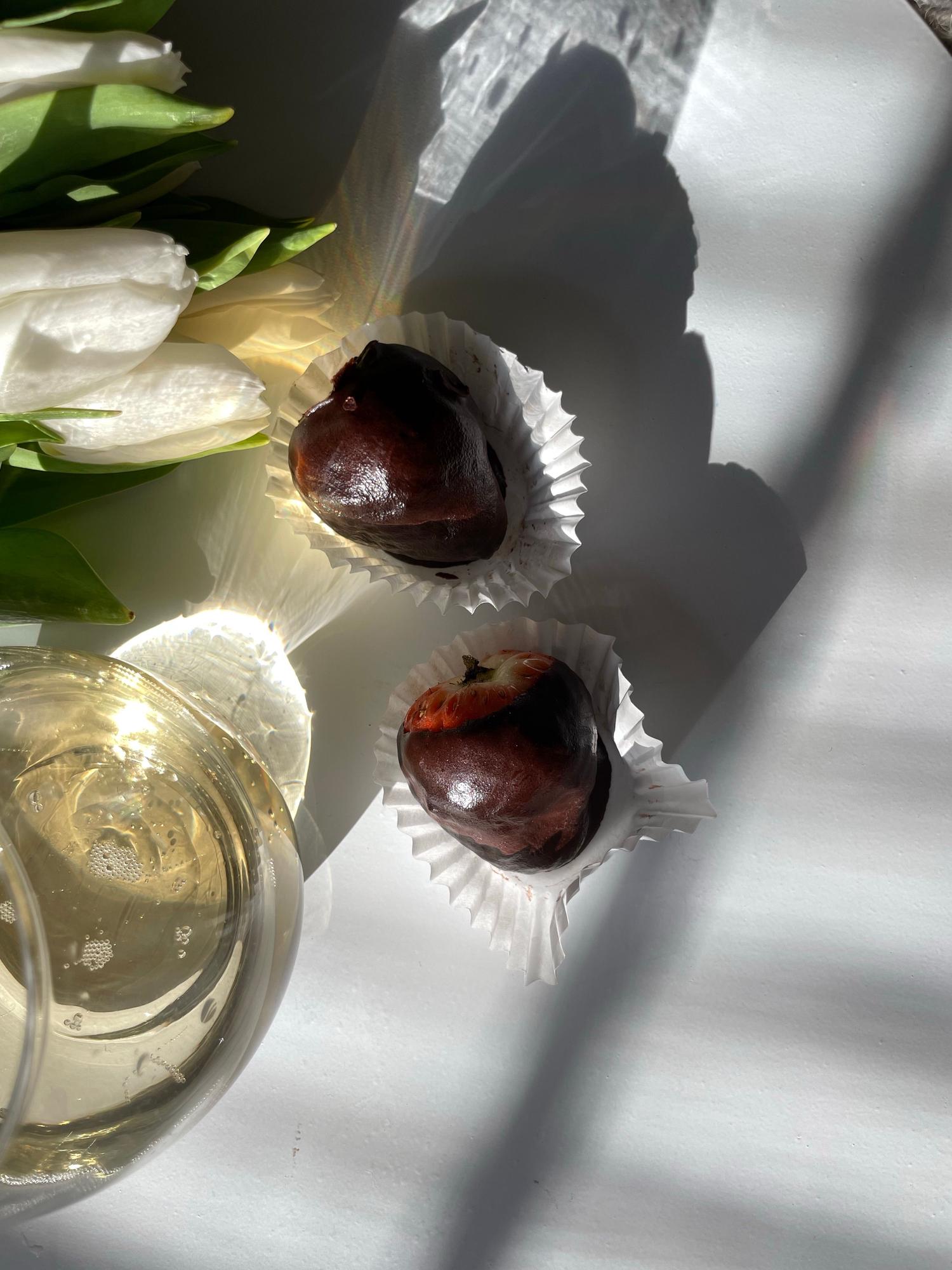 Two dark chocolate-covered strawberries in white liners sit beside white tulips and a sparkling wine glass, illuminated by bright sunlight and dramatic shadows.