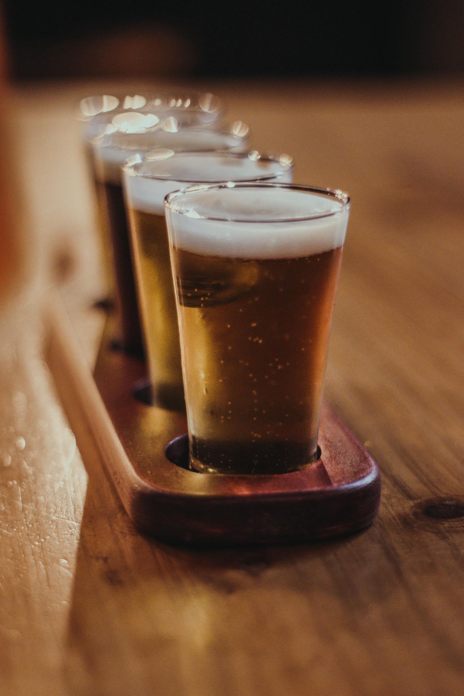 A wooden paddle holds a flight of four small beer glasses, containing different shades of beer with foamy heads, on a wooden table.