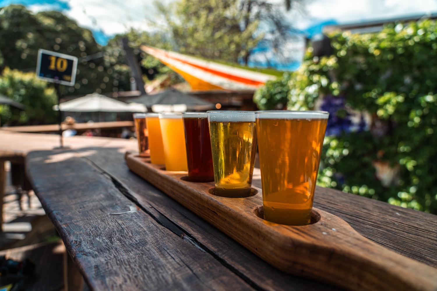 A wooden flight board holds five glasses of craft beer, varying in color from light golden to dark amber, on a rustic outdoor table. A blurred background reveals a sunny patio setting with green foliage and umbrellas.