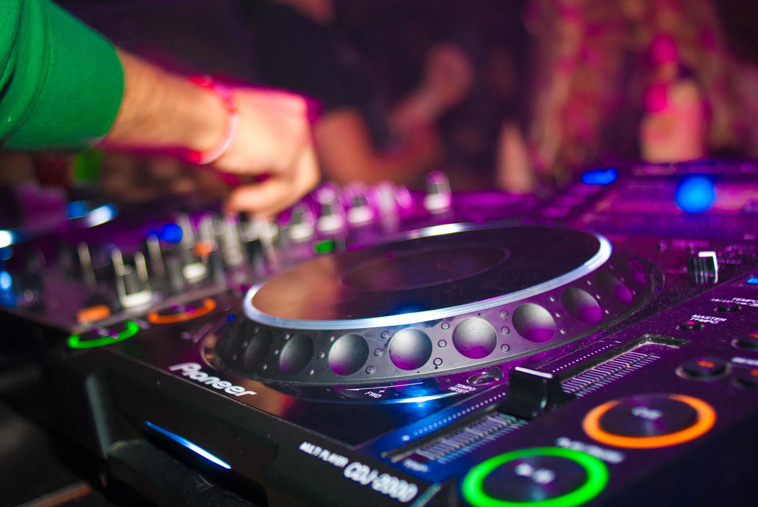 A DJ's hand adjusts controls on a Pioneer mixing console, illuminated by vibrant purple, blue, green, and orange lights in a dark club setting.